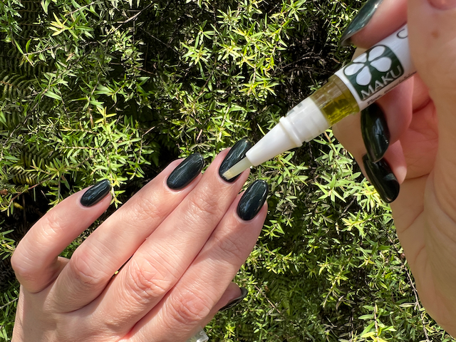 A close up of applying cuticle oil from a Maku Cuticle Oil pen to manicured nails. The nails are painted a shiny green, and the oil is being applied with the right hand to the left. The background is a mānuka bush's green small pointy leaves.
