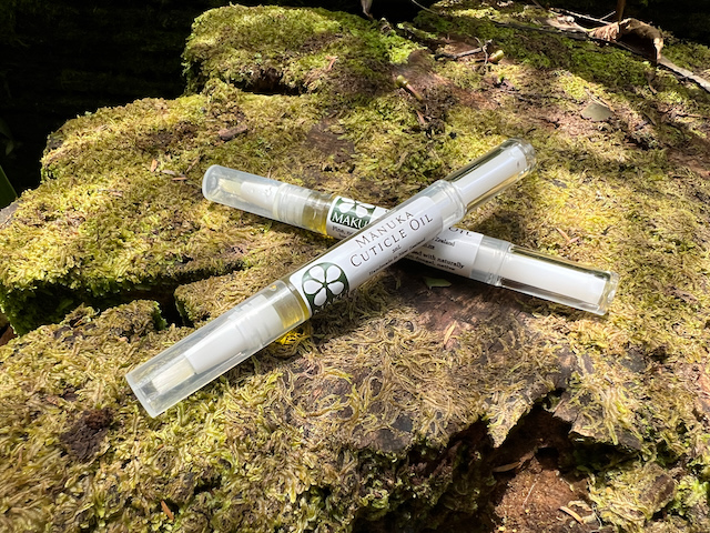 Two pens of cuticle oil displayed on a mossy stump. The oil is green and the pens are a transparent cylinder with a brush applicator.